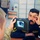 A military or security personnel member in camouflage uniform is conducting an iris scan on a man wearing a dark blue uniform with Arabic text and 'Protection Force' insignia. The handheld biometric device displays a close-up iris image on its screen. This appears to be a biometric enrollment or identity verification procedure in an office or administrative setting.