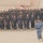A large formation of uniformed personnel standing at attention in an arid, desert-like outdoor setting. A single officer in a distinctly different camouflage uniform walks in front of the assembled group, suggesting an inspection or parade. A red and white flag or pennant is visible in the background among the formation.