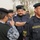 The image shows a group of uniformed personnel identified as members of an 'Oil Protection Force,' based on the visible patch on the right foreground figure's uniform. The scene appears to be an outdoor assembly or inspection, with multiple personnel in dark navy/black uniforms and black berets. A person in camouflage clothing appears to be interacting with or inspecting the uniformed personnel.