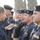 A group of uniformed personnel, appearing to be Iraqi police officers based on visible insignia and flag patches, are standing in a line formation outdoors. A civilian or plainclothes individual on the left is writing on a notepad, possibly conducting an inspection, roll call, or interview. The setting appears to be an outdoor compound with trees visible in the background.