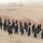 A large group of uniformed personnel in black uniforms and tan/desert boots are marching in formation along a paved road. They appear to be engaged in a training or drill exercise in an arid, desert-like environment. The group consists of approximately 30-40 individuals wearing matching insignia patches on their sleeves.