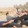 A large group of individuals in dark athletic/uniform clothing are performing paired sit-up exercises in a long row on a paved surface. The setting appears to be an outdoor training facility or military/police training ground in an arid environment. An instructor or supervisor is visible standing in the background overseeing the exercise.