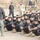 A large group of uniformed personnel wearing matching dark/black uniforms and black berets are seated on the ground in an outdoor setting, apparently receiving instruction or a briefing. A bald instructor in civilian-style tactical clothing stands facing the group, appearing to address them. Several standing supervisors or observers are visible in the background.