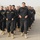 A large group of uniformed personnel in black uniforms and tan boots are marching in formation along a paved road. An individual wearing a peaked cap walks alongside the formation, likely serving as an instructor or supervisor. The setting appears to be a training facility or base, visible from the concrete barrier walls and arid landscape in the background.