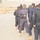 A uniformed instructor or officer is directing a large group of trainees or recruits during what appears to be an outdoor drill or formation exercise. The personnel are dressed in dark navy/blue uniforms with black belts and tan boots, suggesting a police or paramilitary training context. The setting is an arid, open outdoor area with sparse vegetation and hazy atmospheric conditions.