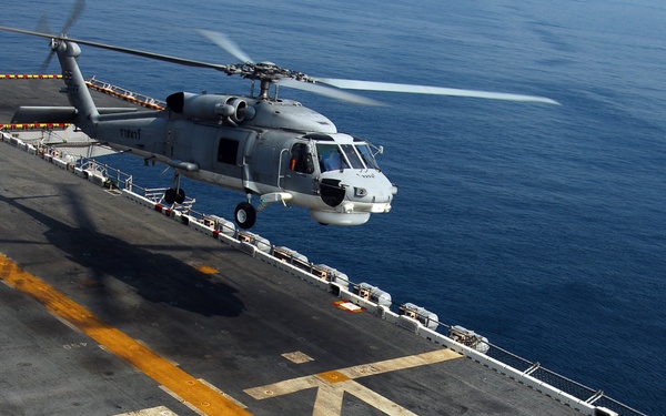 An aerial view of a naval vessel's flight deck showing a military helicopter in the process of landing or hovering. Two flight deck crew members in yellow jerseys are visible on the deck, with one appearing to signal the helicopter. The ocean is visible in the background, and the deck features standard flight deck markings including yellow lines and numbered designations.