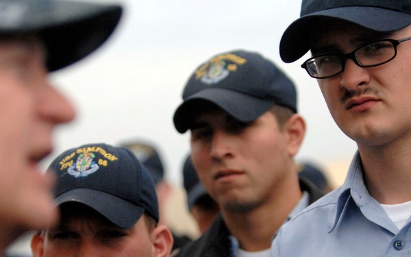 The image shows a group of U.S. Navy sailors in light blue utility uniforms and dark navy ball caps, appearing to be at attention or receiving a briefing. At least two sailors have visible name tapes and 'U.S. NAVY' branch labels on their shirts. The sailor on the right is identified as 'SPRADLING' and wears black-framed glasses.