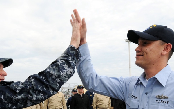 Two U.S. Navy personnel are giving each other a high-five on what appears to be the deck of a naval vessel. The individual on the left is wearing Navy Working Uniform (NWU) digital camouflage, while the person on the right is in a light blue Navy uniform with a name tag reading 'RIDAN' and a Petty Officer insignia. Several other Navy personnel in khaki and dark uniforms are visible in the background.