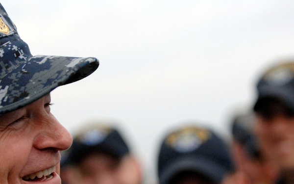 A senior U.S. Navy officer in the foreground is smiling, wearing a Navy Working Uniform (NWU) digital camouflage pattern with a gold insignia on his cap. Behind him, several other naval personnel are visible in soft focus, wearing dark navy ball caps with insignia. The setting appears to be an outdoor military environment.