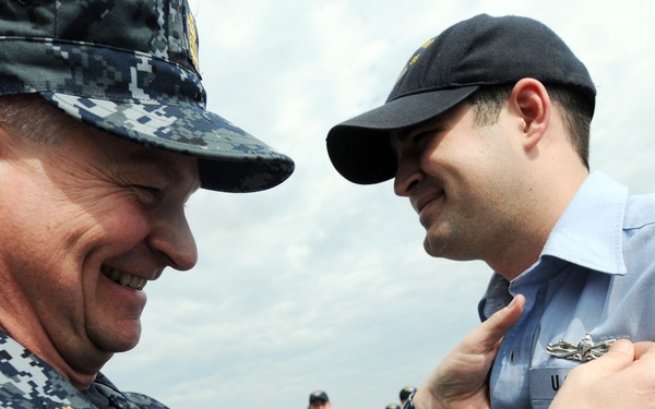 A senior U.S. Navy officer in Navy Working Uniform (NWU) is pinning a badge or insignia onto a junior sailor's uniform during what appears to be a formal recognition or qualification ceremony. Both individuals are smiling, suggesting a positive and celebratory occasion. The setting appears to be an outdoor naval environment, likely a ship's deck, with other personnel visible in the background.