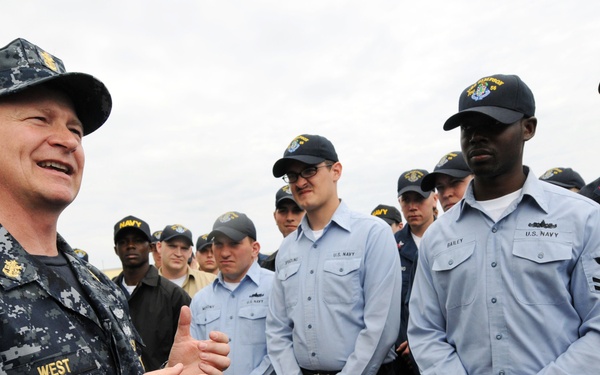 A senior U.S. Navy officer with the name tape 'WEST' on his NWU (Navy Working Uniform) Type I digital camouflage is addressing a group of enlisted sailors. The sailors are wearing a mix of NWU Type I (dark blue digital camo) and Navy Working Uniform blue shirts, all wearing Navy ball caps. The setting appears to be an outdoor naval environment, possibly a ship's deck or pier.