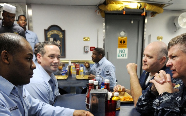 A group of U.S. Navy personnel are seated at tables in what appears to be a ship's mess hall or dining facility. A senior officer in Navy Working Uniform (NWU) digital camouflage pattern is engaged in conversation with enlisted sailors wearing light blue utility uniforms. The setting suggests an informal meeting or meal engagement aboard a naval vessel.