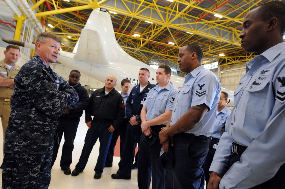 A senior U.S. Navy officer in Navy Working Uniform (NWU) camouflage is addressing a group of enlisted sailors in a military aircraft hangar. The sailors are wearing a mix of NWU camouflage, service khaki, and Navy blue working uniforms. A large white military aircraft, likely a P-8 Poseidon or P-3 Orion, is visible in the background.