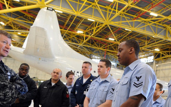 A senior U.S. Navy officer in Navy Working Uniform (NWU) camouflage is addressing a group of enlisted sailors in a military aircraft hangar. The sailors are wearing a mix of NWU camouflage, service khaki, and Navy blue working uniforms. A large white military aircraft, likely a P-8 Poseidon or P-3 Orion, is visible in the background.