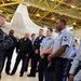A senior U.S. Navy officer in Navy Working Uniform (NWU) camouflage is addressing a group of enlisted sailors in a military aircraft hangar. The sailors are wearing a mix of NWU camouflage, service khaki, and Navy blue working uniforms. A large white military aircraft, likely a P-8 Poseidon or P-3 Orion, is visible in the background.