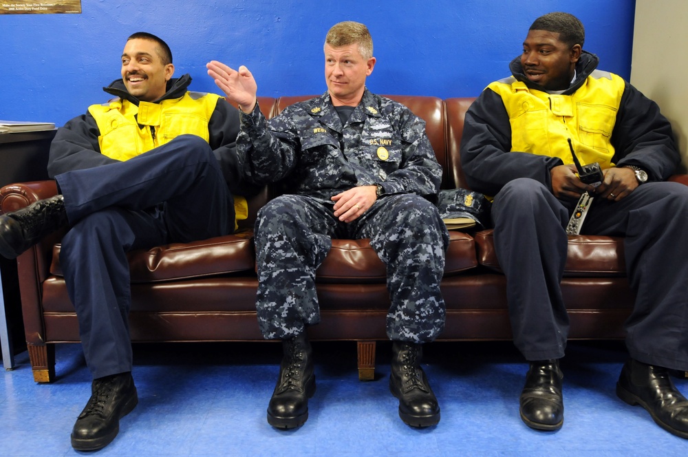 Three men are seated on a brown leather couch against a blue wall. The center individual is wearing a U.S. Navy NWU (Navy Working Uniform) with visible rank insignia and a name tape reading 'WEST', while the two flanking individuals wear yellow high-visibility safety vests over dark clothing. They appear to be engaged in casual conversation, with the center person gesturing with his hand.