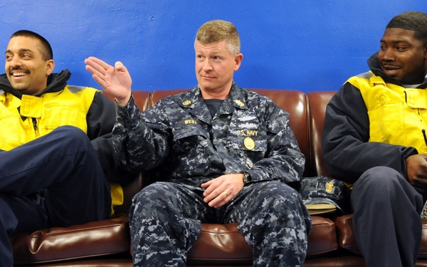 Three men are seated on a brown leather couch against a blue wall. The center individual is wearing a U.S. Navy NWU (Navy Working Uniform) with visible rank insignia and a name tape reading 'WEST', while the two flanking individuals wear yellow high-visibility safety vests over dark clothing. They appear to be engaged in casual conversation, with the center person gesturing with his hand.