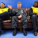Three men are seated on a brown leather couch against a blue wall. The center individual is wearing a U.S. Navy NWU (Navy Working Uniform) with visible rank insignia and a name tape reading 'WEST', while the two flanking individuals wear yellow high-visibility safety vests over dark clothing. They appear to be engaged in casual conversation, with the center person gesturing with his hand.