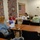 A sewing class or workshop is taking place in a room decorated with patriotic quilts and American flag-themed textile art. Three students are seated at tables with sewing machines while an instructor stands and observes. The setting appears to be a community or military-affiliated facility based on the Department of the Army seal visible on one of the quilts.