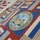 A patriotic patchwork quilt featuring the official seal of the United States Department of the Army as its central medallion. The quilt incorporates red, white, blue, and tan fabrics with American flag motifs, stars, and stripes in a traditional block pattern. The Army seal displays the founding year 1775 and the motto 'This We'll Defend.'