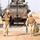 Two soldiers in desert camouflage uniforms and body armor walk forward on a dirt road in an arid environment. A large tan military armored vehicle (appears to be an MRAP or similar heavy tactical vehicle) is visible in the background. Additional personnel are visible in the far background near the vehicle.