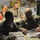 Military personnel in combat uniforms are gathered inside what appears to be a forward operating base tent or temporary facility. Two soldiers in the foreground are reviewing documents in a binder, likely maps or operational materials. Additional soldiers are visible in the background near food supplies and a chest freezer.