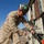 A military service member in desert camouflage uniform is working on an electrical or telecommunications panel/junction box outdoors. The individual is wearing a hard hat with a green sticker and safety glasses, suggesting adherence to safety protocols. The setting appears to be a forward operating base or military installation in an arid environment.