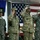 Four military personnel in uniform are standing at attention and saluting during a formal ceremony. They are positioned in front of a large American flag and what appears to be a US Air Force flag on the left. Name tapes visible on uniforms include 'McMahon', 'Kornatz', and 'Morgan'.