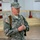 A U.S. Army Colonel in Army Combat Uniform (ACU) pattern is standing and appears to be speaking or briefing. The name tape reads 'MCMAHON' and rank insignia (eagle) is visible on both the patrol cap and chest. He is carrying a brown leather strap bag and appears to be in an indoor facility.