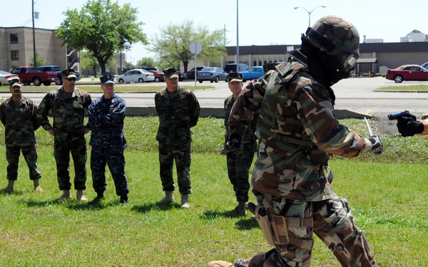 A military or law enforcement training exercise is taking place outdoors on what appears to be a base or installation. Two participants in full protective gear and helmets are engaged in a close-quarters combat or apprehension drill, with one running and one pointing a training weapon. A group of approximately seven uniformed observers in mixed military attire watch from the sideline.