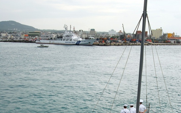 Naval personnel in white dress uniforms are standing at attention along the bow rail of a warship, performing a 'manning the rails' ceremonial formation. A Japan Coast Guard vessel (PL63) is visible in the background across the harbor. The scene appears to be a port arrival or departure ceremony at a Japanese harbor.