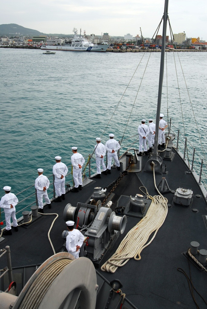 Naval personnel in white dress uniforms are standing at attention along the bow rail of a warship, performing a 'manning the rails' ceremonial formation. A Japan Coast Guard vessel (PL63) is visible in the background across the harbor. The scene appears to be a port arrival or departure ceremony at a Japanese harbor.