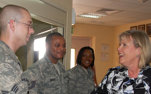 A civilian woman in a floral blouse is shaking hands with a U.S. Air Force staff sergeant in ABU (Airman Battle Uniform) camouflage. The setting appears to be an indoor military facility or office, with additional uniformed personnel visible in the background. The interaction appears to be a formal greeting or recognition event.