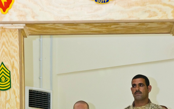 A military ceremony or memorial service featuring U.S. Army and what appears to be Iraqi Army soldiers standing at attention. Red, white, and blue candles are visible in the foreground, suggesting a formal commemorative event. The setting appears to be an indoor military facility decorated with U.S. Army sergeant major rank insignia on a plywood wall.