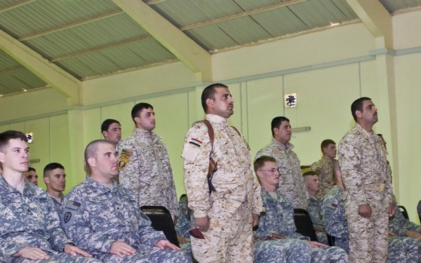 A group of military personnel from multiple nations are assembled in a large indoor facility, likely during a formal ceremony or joint meeting. Soldiers are wearing at least three distinct camouflage uniform patterns, suggesting a multinational military gathering. Some personnel are seated while others stand in formation.