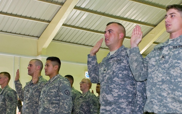 A group of U.S. Army soldiers in ACU (Army Combat Uniform) digital camouflage pattern are standing in a line with their right hands raised, appearing to take an oath or re-enlistment ceremony. The event is taking place indoors in what appears to be a military facility or chapel with a corrugated metal roof. Additional soldiers are seated in the background observing the ceremony.