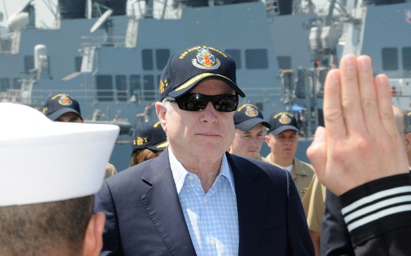 A civilian official wearing a dark navy cap and sunglasses stands on the deck of a naval vessel, holding a blue folder, while a sailor in dress whites raises their right hand in what appears to be an oath or re-enlistment ceremony. Multiple uniformed Navy personnel in both dress whites and working khaki uniforms are visible in the background. Large naval warships are docked in the background.