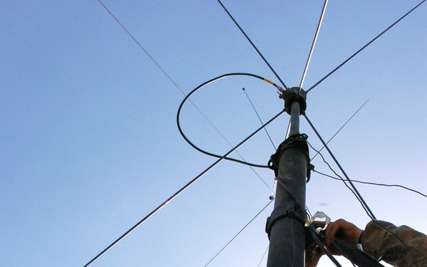 Two soldiers in US Army ACU (Army Combat Uniform) digital camouflage are erecting or adjusting a military communications antenna mast. The image is taken from a low angle looking upward, showing the soldiers working on the upper section of the mast with multiple antenna elements and guy wires visible against a blue sky. The antenna assembly at the top appears to include a loop element and multiple radial wire elements.