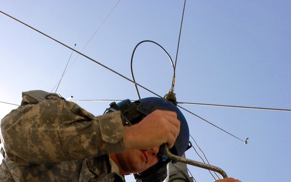 A U.S. Army soldier in Army Combat Uniform (ACU) digital camouflage pattern is working at the top of what appears to be a communications antenna mast or tower structure. The soldier is using a hand tool, likely a wrench or ratchet, to tighten or adjust hardware on the mast. A second soldier in matching ACU is visible below, appearing to stabilize or assist with the structure.