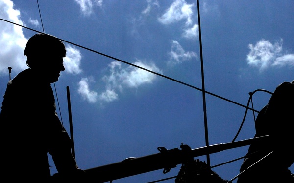 A dramatic silhouette photograph showing two helmeted figures working with equipment against a bright blue sky with scattered white clouds. The strong backlighting renders both figures and surrounding structures in near-total silhouette, creating a high-contrast composition. The figures appear to be handling a long pole or beam amid a network of cables and structural elements.