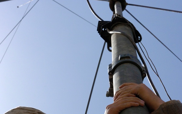 Two soldiers in US Army ACU (Army Combat Uniform) digital camouflage are working together to assemble or secure a military antenna mast. One soldier wearing a combat helmet is using a small tool, likely a pin or locking mechanism, to secure a joint connection on the mast. The photo is taken from a low angle looking upward, with guy wires visible extending from the top of the mast against a clear blue sky.