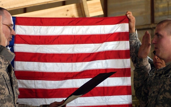 Two U.S. Army soldiers in ACU (Army Combat Uniform) are conducting a military oath ceremony, likely a reenlistment or promotion, in what appears to be a forward operating base or field environment. The soldier on the left is reading from a document while the soldier on the right has his right hand raised. An American flag is displayed as a backdrop behind them.