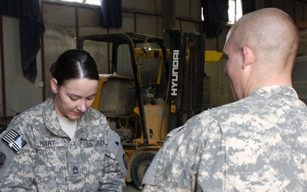 Two U.S. Army soldiers in ACU (Army Combat Uniform) are reviewing and signing paperwork in what appears to be a maintenance or logistics facility. The female soldier, identified by her name tape as 'HART,' is writing on a document held in a folder. A Hyundai forklift is visible in the background of the industrial workspace.