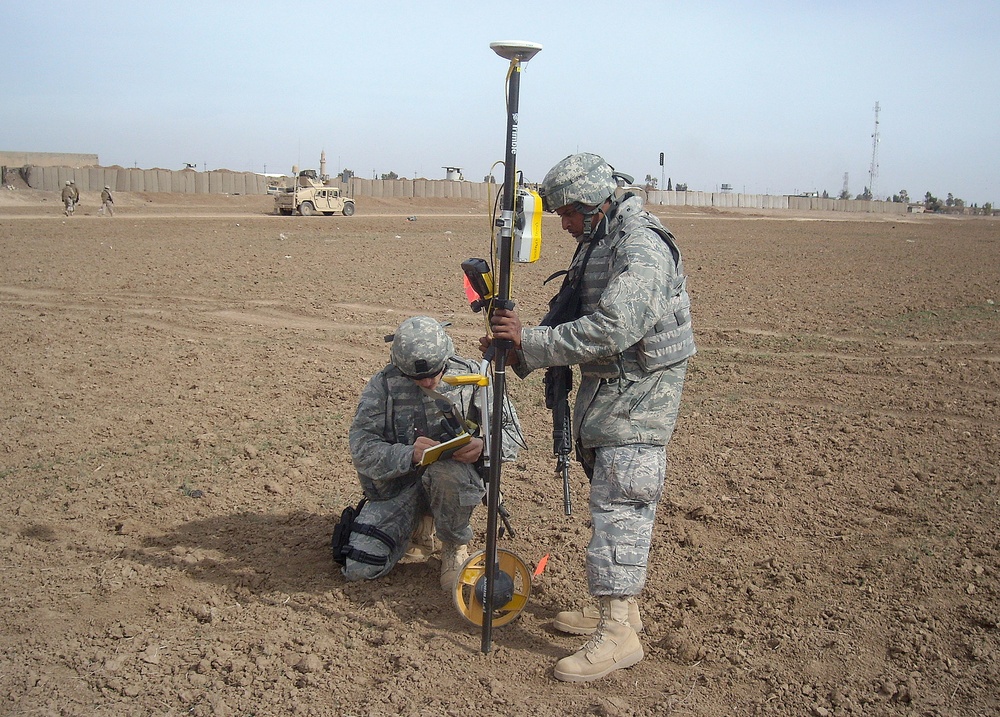 Two military personnel in US Army/Air Force ACU/ABU camouflage uniforms are conducting a land survey operation in an arid, open field. One soldier is crouching and recording data on a clipboard while the other stands holding a Trimble GPS survey pole with a measuring wheel attached. The background shows a military compound with concrete barriers (T-walls/HESCO barriers), Humvees, and additional personnel.