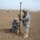 Two military personnel in US Army/Air Force ACU/ABU camouflage uniforms are conducting a land survey operation in an arid, open field. One soldier is crouching and recording data on a clipboard while the other stands holding a Trimble GPS survey pole with a measuring wheel attached. The background shows a military compound with concrete barriers (T-walls/HESCO barriers), Humvees, and additional personnel.