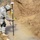 A U.S. Army soldier in ACU (Army Combat Uniform) is conducting what appears to be a survey or geospatial measurement operation in a muddy, water-filled excavation or trench. The soldier is operating a GPS/GNSS survey rod with a dome antenna on top and a handheld data collector device. The environment shows exposed earth walls and shallow muddy water, suggesting a field engineering or reconnaissance task.