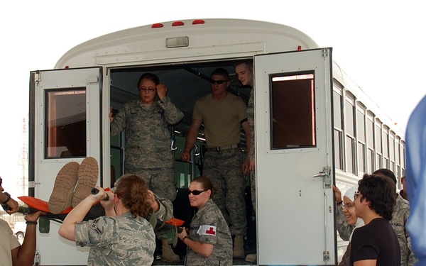 Military personnel in US Air Force camouflage uniforms are unloading a patient on a stretcher from the rear doors of a white bus. At least two personnel wearing red cross armbands are visible, indicating medical staff involvement in what appears to be a patient transfer or medical evacuation exercise. A civilian woman wearing a blue hijab is visible in the foreground right.