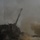 The image shows a self-propelled artillery system firing or having just fired, surrounded by dense smoke and dust clouds. The vehicle's long gun barrel is elevated at an angle, consistent with an artillery firing position. Visibility is heavily obscured by discharge smoke and disturbed dust.