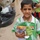 A young boy, approximately 6-8 years old, stands holding a packaged item that includes green gloves and what appears to be toy soldiers or figurines, with Arabic text visible on the packaging. A U.S. military soldier in ACU (Army Combat Uniform) is partially visible on the left side of the frame. The scene appears to be a humanitarian aid or community outreach distribution event.
