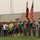 A ceremonial opening of a soccer tournament at a stadium, featuring U.S. military personnel in ACU (Army Combat Uniform) holding the American flag and what appears to be the Iraqi flag alongside local civilian soccer players in various colored jerseys. The event appears to be a joint U.S.-Iraqi community sports event, likely during the Iraq War era based on uniforms and context. The stadium seating is largely empty with a hazy, overcast sky visible in the background.